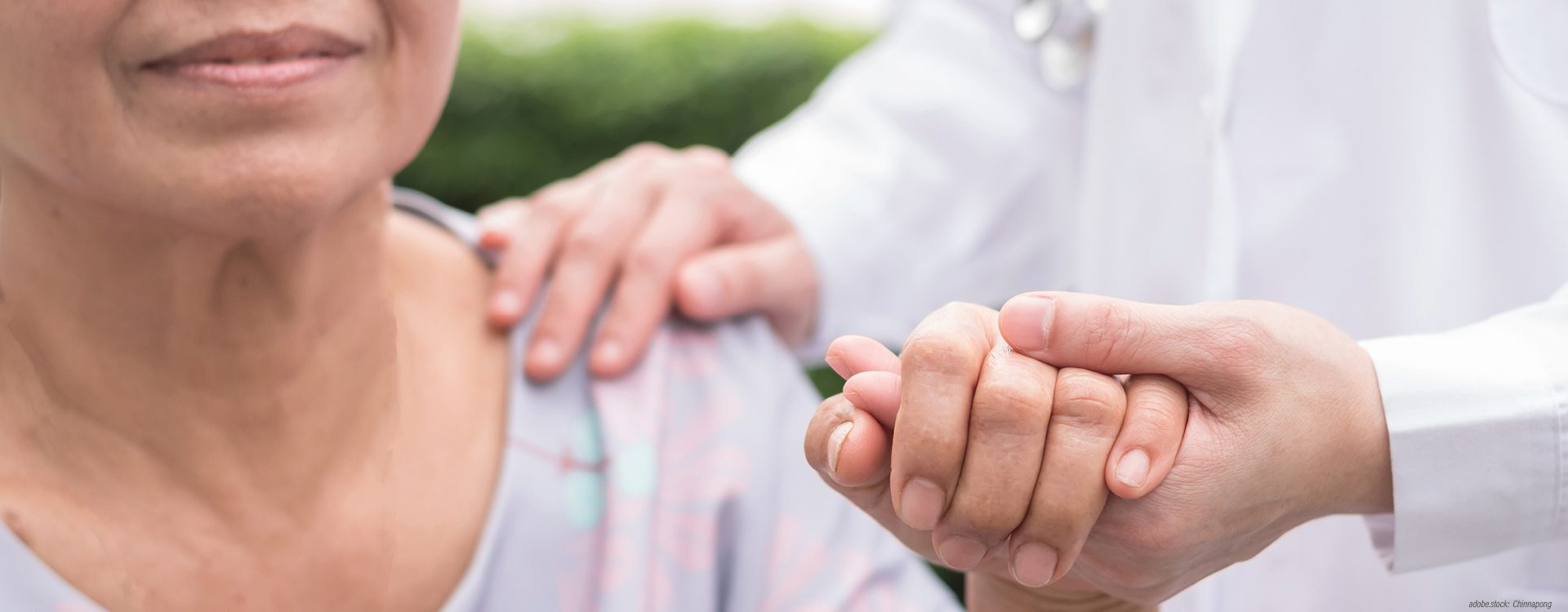 zwei ausschnittsweise dargestellte Personen: eine Person in weißer Krankenhausarbeitskleidung hat die rechte Hand auf die Schulter der anderen Person gelegt, mit ihrer linken Hand hält sie die linke Hand der Patientin