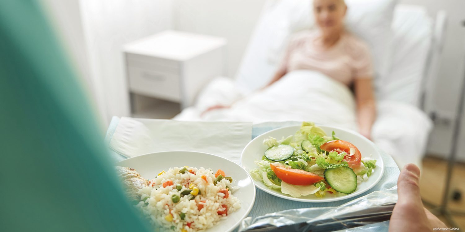 Tablett mit zwei gefüllten Tellern wird zu einer unscharf dargestellten, im Bett liegenden Patientin gebracht