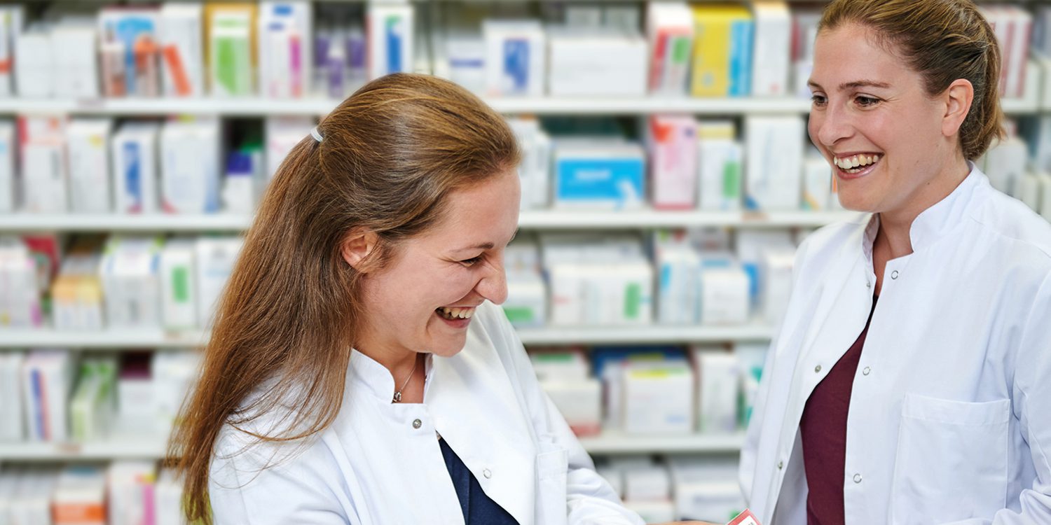 zwei freundliche weibliche Personen in Krankenhausarbeitskleidung vor einem mit Medikamentenpackungen gefüllten Regal in einer Apotheke