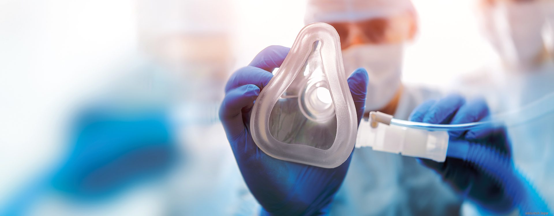 unscharf dargestellte Person in OP-Arbeitskleidung zeigt eine Beatmungsmaske