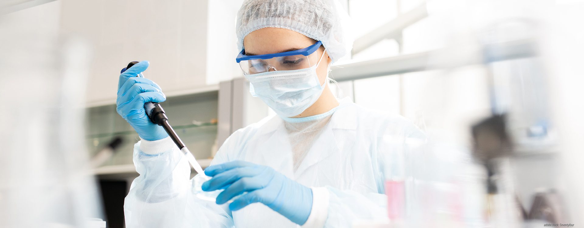 Eine Laborfachkraft in Arbeitsschutzkleidung gibt mit einer Pipette ein Flüssigkeit in ein flaches Glasschälchen.