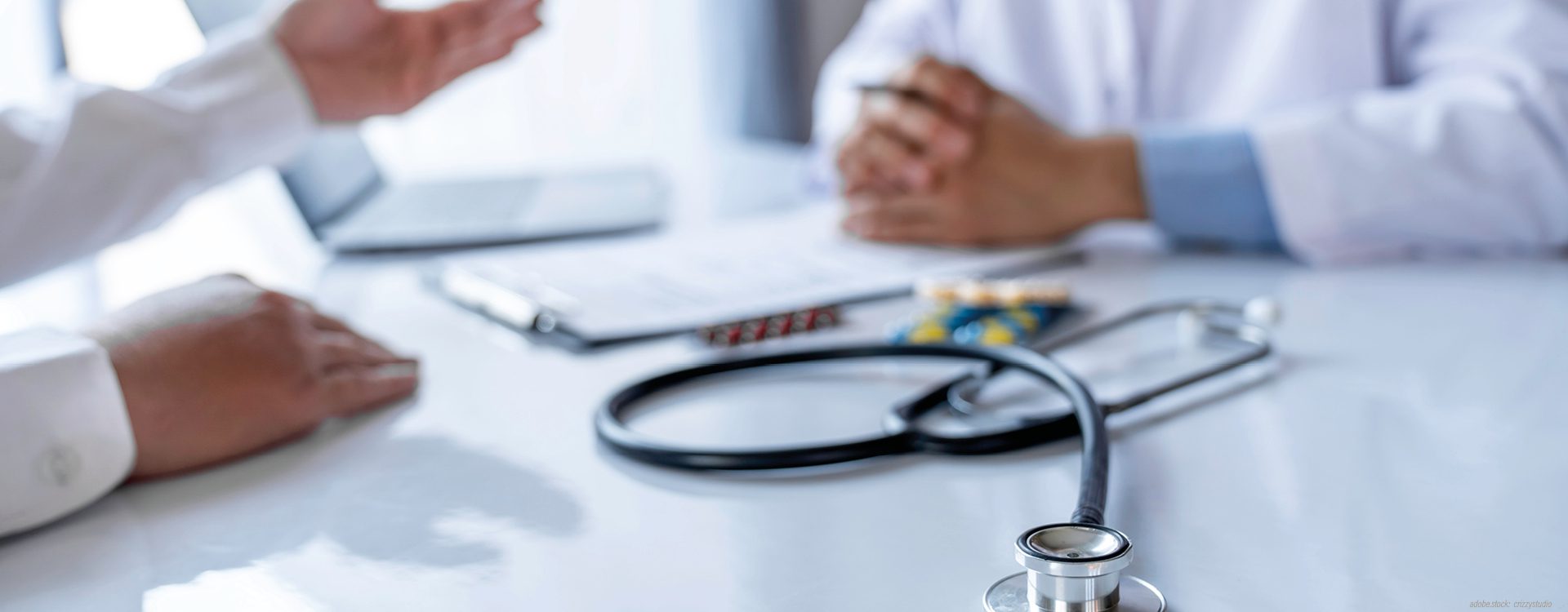 Zwei Personen sitzen sich an einem Tisch gegenüber und unterhalten sich. Zu sehen sind nur ihre Hände. Auf dem Tisch liegt ein Stethoskop.