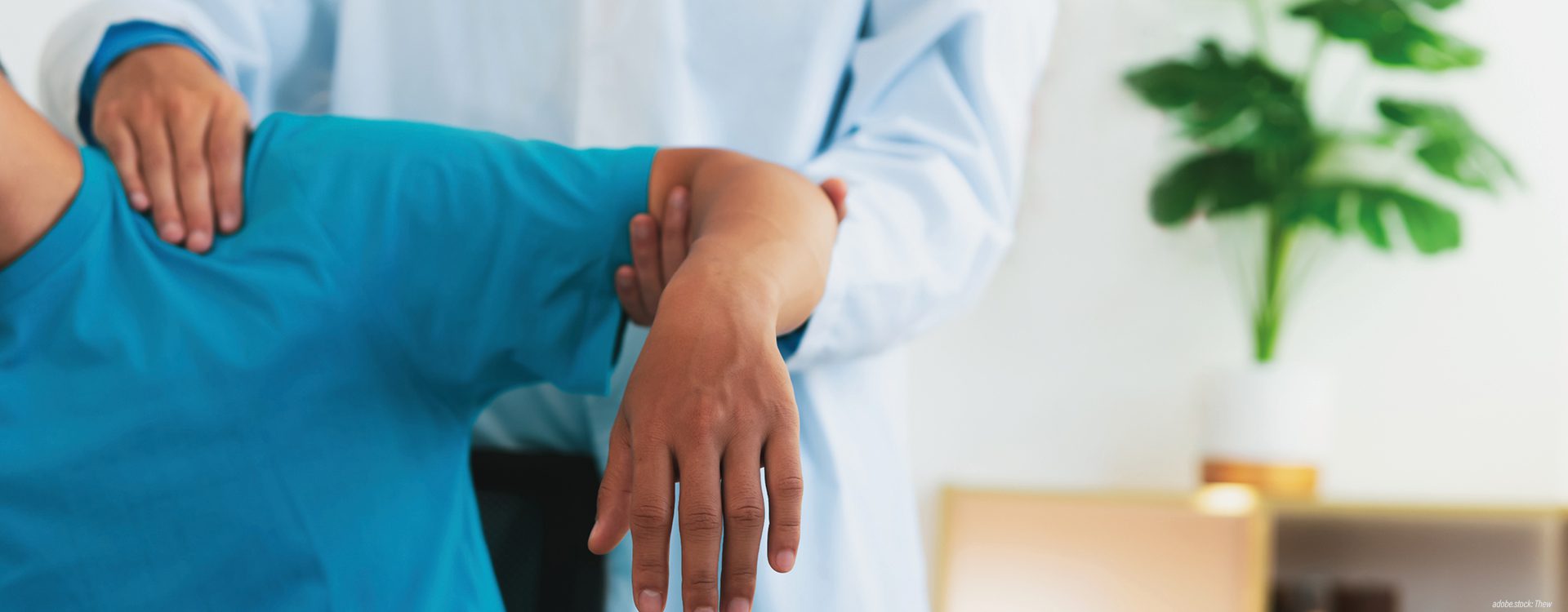 Eine nur ausschnittsweise abgebildete Person in Krankenhausarbeitskleidung untersucht die Schulter einer Person.