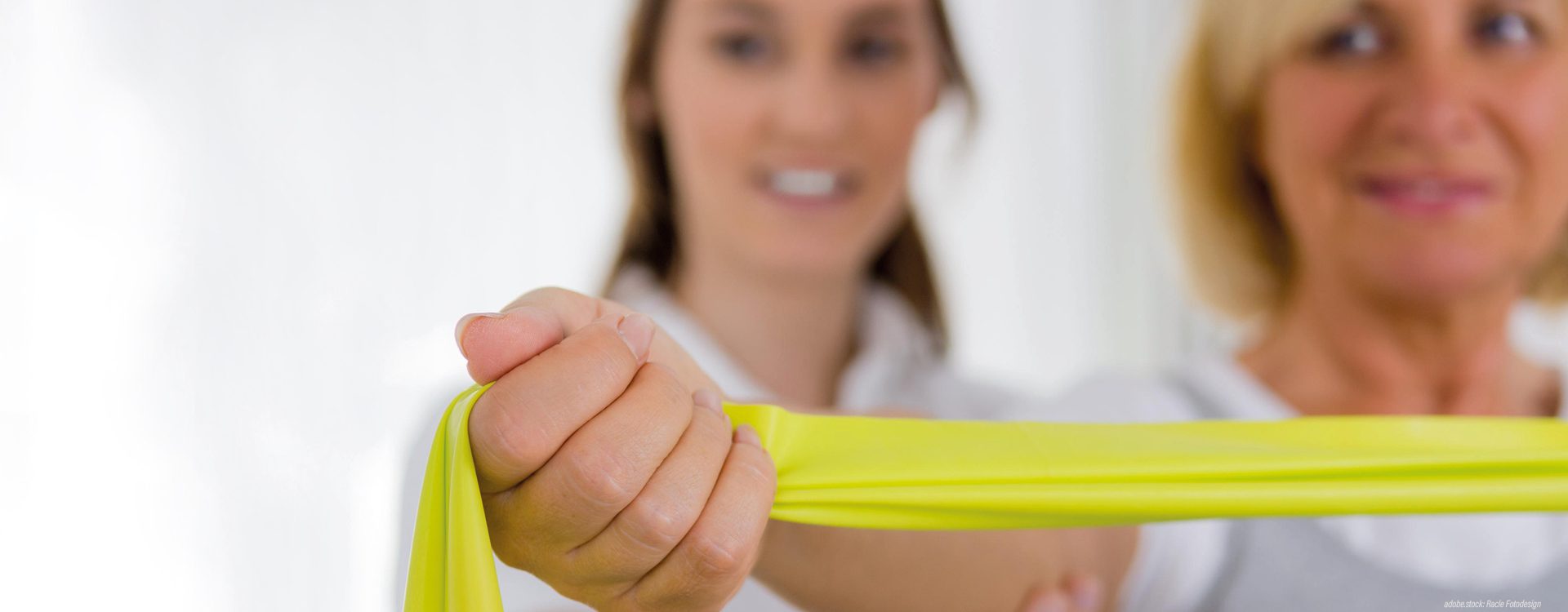 Zwei lächelnde, unscharf frontal abgebildete weibliche Personen. Im Vordergrund eine Patientin mit gelbem Stretchband, die Physiotherapeutin steht hinter ihr und unterstützt sie