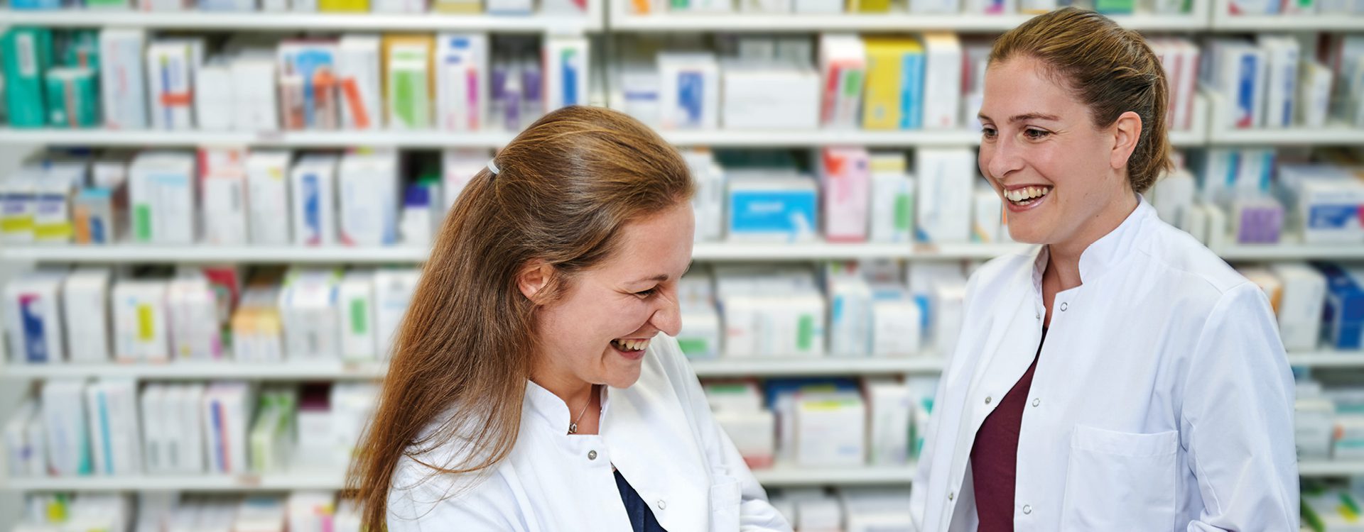 zwei freundliche weibliche Personen in Krankenhausarbeitskleidung vor einem mit Medikamentenpackungen gefüllten Regal in einer Apotheke