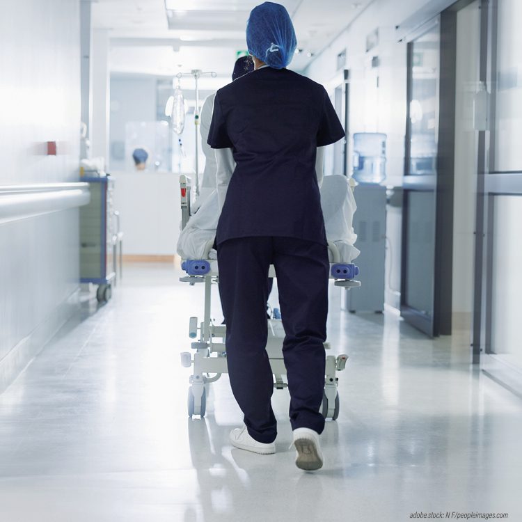 Person in blauer Krankenhausarbeitskleidung schiebt eilig eine Trage mit einer liegenden Person einen Krankenhausflur entlang